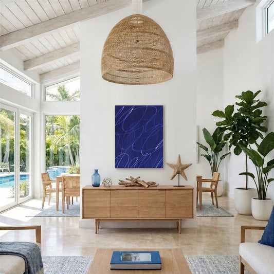 A modern coastal-style living room featuring a prominent blue abstract wall art print with white lines, styled above a light wood credenza. The room has high ceilings with exposed wood beams, a large woven wicker pendant light, and is surrounded by green plants in white planters.